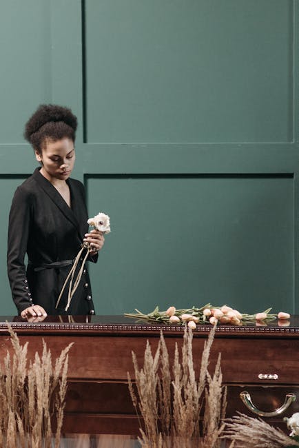 A woman with dark curly hair styled in an elegant updo, dressed in a black satin blazer, stands beside a polished wooden table holding a small, delicate white flower with soft pink accents, possibly a