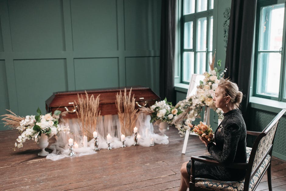 A woman seated indoors on a patterned armchair holding a floral bouquet, facing a large floral arrangement atop a white easel. The arrangement features white and blush roses, peonies, and greenery, wi
