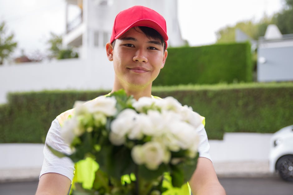 A cheerful delivery person wearing a red cap and a light-colored uniform is standing outdoors on a residential street, holding a bouquet of fresh white flowers with green foliage. The bouquet appears 