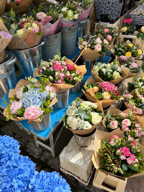 A display of fresh flower bouquets arranged in metal buckets and brown paper wraps on a blue table at a florist shop. The bouquets feature a variety of flowers including pink roses, white roses, light