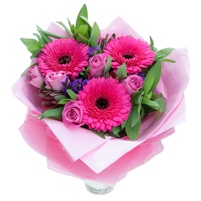 I see this as a bouquet that balances confident colour with gentle intimacy: three fuchsia-pink Gerbera daisies stand as lively focal points, their flat, layered petals and deep dark centers creating bold structure, while surrounding lavender-pink roses offer softer, swirling forms that invite closer inspection. The roses are in that pleasing, early stage where their petals are still snug, suggesting freshness and a slow reveal of scent. Tiny sprays of purple statice tuck into gaps between the larger flowers, providing a slightly papery texture and a cool violet accent that gives the eye places to rest. The foliage - broad, glossy leaves similar to salal or eucalyptus - forms a supportive, textured frame, imparting a crisp green note and a subtle citrus-wood fragrance when handled. The bouquet is wrapped with considered craftsmanship: an inner layer of pale pink matte tissue creates a gentle visual base, and a translucent, slightly crinkled fuchsia-pink cellophane overlays it with a soft shimmer, the folds neatly arranged to hold the blooms securely. You can glimpse the top rim of a clear glass vase at the base, suggesting the piece is intended for immediate display on a mantel or kitchen table. In the soft morning light common to our neighbourhood, the composition reads as a warm, intimate gift - suitable for celebrating small joys, anniversaries, or simply to brighten someone's day after a walk through nearby Crystal Palace Park or a coffee on Westow Street.